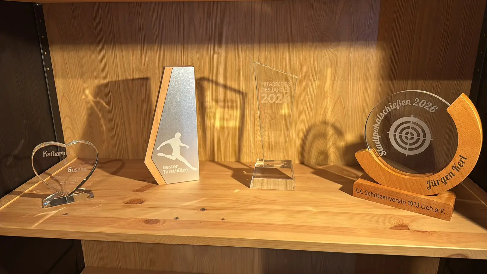 Vier gravierte Muster-Trophäen nebeneinander auf einem Holzregal im Laden: ein Herz aus Glas, ein Holzpokal mit Aluminiumfront und Fußballer-Motiv, ein Glaspokal „Mitarbeiter des Jahres 2026“ und ein runder Glaspokal im Holzbogen für ein Stadtpokalschießen